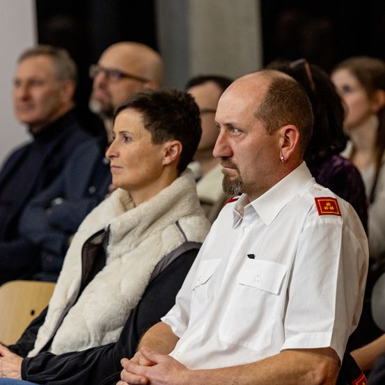 Eine Gruppe von Menschen sitzt in einem Raum, ein Mann in weißer Uniform und eine Frau in einer weißen Pelzweste. Sie scheinen an einer formellen Veranstaltung teilzunehmen.