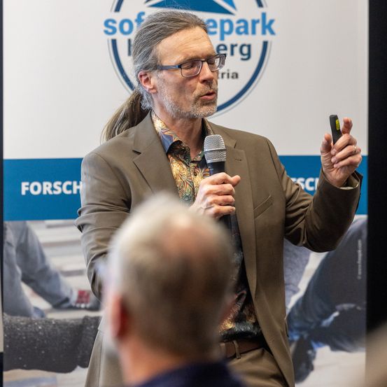 Ein Mann mit einem Pferdeschwanz hält ein Mikrofon und hält eine Präsentation vor einem Publikum. Hinter ihm ist ein Banner mit dem Text 'sofepark berg'.