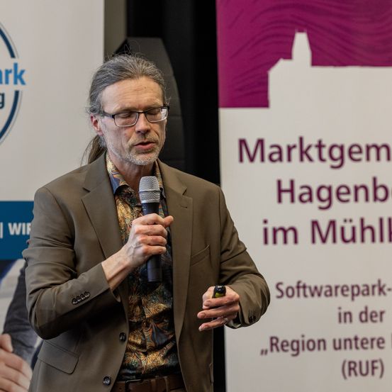Ein Mann mit Brille und einem braunen Anzug spricht in ein Mikrofon. Hinter ihm befindet sich ein violetter und weißer Schild mit deutschem Text.
