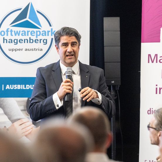 Ein Mann in einem Anzug spricht in ein Mikrofon auf einer Konferenz vor einem Softwarepark Hagenberg-Banner.