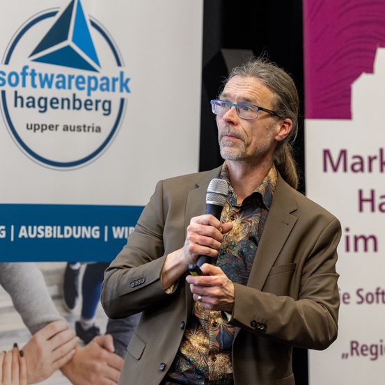 Ein Mann mit Brille steht und spricht in ein Mikrofon vor einem Softwarepark Hagenberg-Schild.