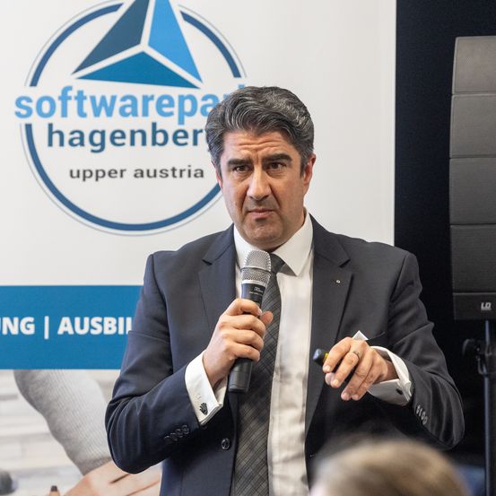 Ein Mann spricht in ein Mikrofon bei einer Veranstaltung vor dem 'softwarepark hagenberg' Banner.