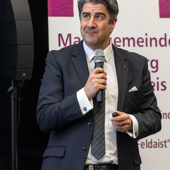 Ein Mann in einem dunklen Anzug und einer Krawatte spricht in ein Mikrofon bei einer Veranstaltung. Er hält einen Stift in der rechten Hand.