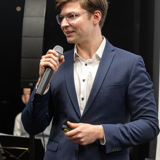 Ein Mann in einem blauen Anzug mit Nadelstreifen hält ein Mikrofon und spricht, während ein anderer Mann in einem weißen Hemd hinter ihm ein Instrument spielt.