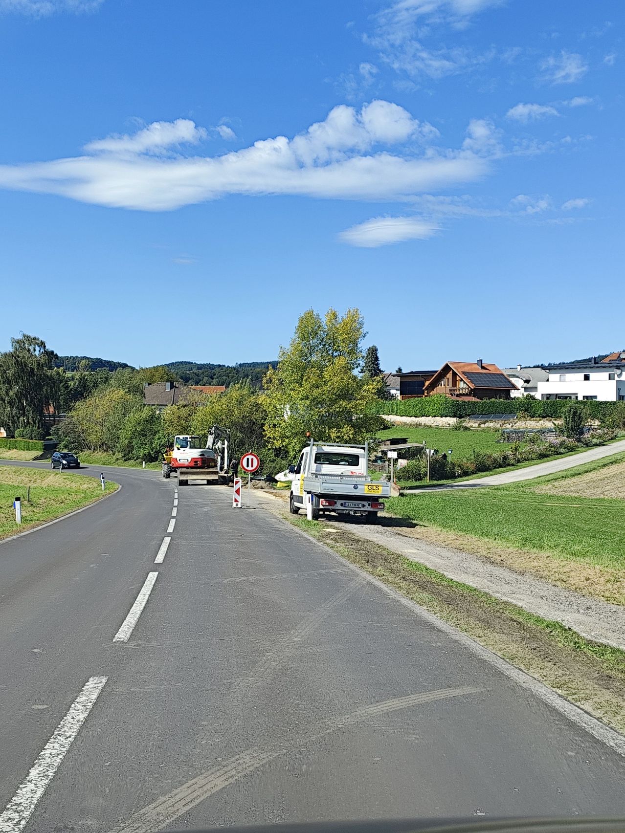 Eine Straße mit geparkten Lastwagen und einem roten Schild ist von Grün umgeben. In der Ferne sind Häuser mit Solarpanelen zu sehen, und ein Auto fährt vorbei. Der Himmel ist klar mit einigen Wolken.