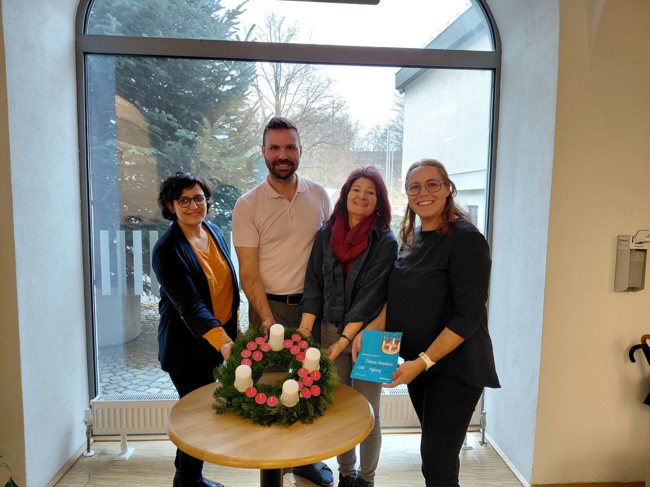 Fünf Menschen stehen in einem Raum, lächelnd, mit einem Kranz aus Kerzen auf einem Tisch, während eine Person ein Zertifikat hält. Hinter ihnen zeigt ein großes Fenster Bäume.