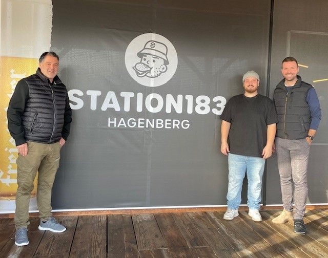 Drei Männer stehen vor einem Schild, das STATION183 HAGENBERG sagt. Sie lächeln und posieren für ein Foto.
