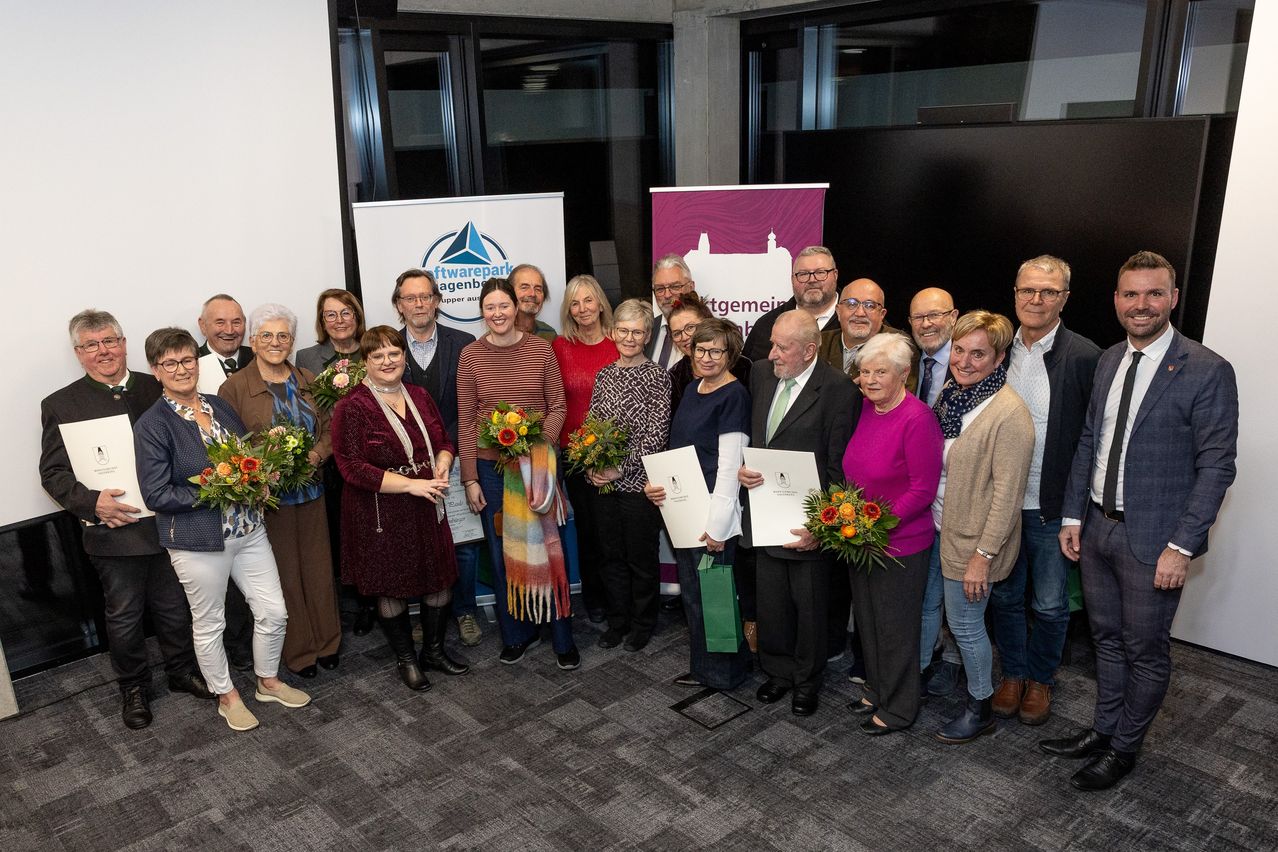 Eine Gruppe von Menschen steht zusammen und hält Blumen und Urkunden. Sie sind in formeller Kleidung gekleidet, und einige tragen Brillen. Ein weißes Banner mit Logo steht hinter ihnen.