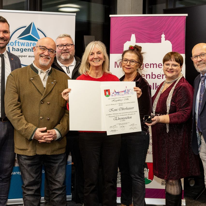 Eine Gruppe von Menschen steht vor einem Hintergrund. Eine Frau hält ein Zertifikat und lächelt. Sie steht zwischen zwei anderen Frauen. Ein Mann auf der linken Seite trägt einen braunen Mantel.