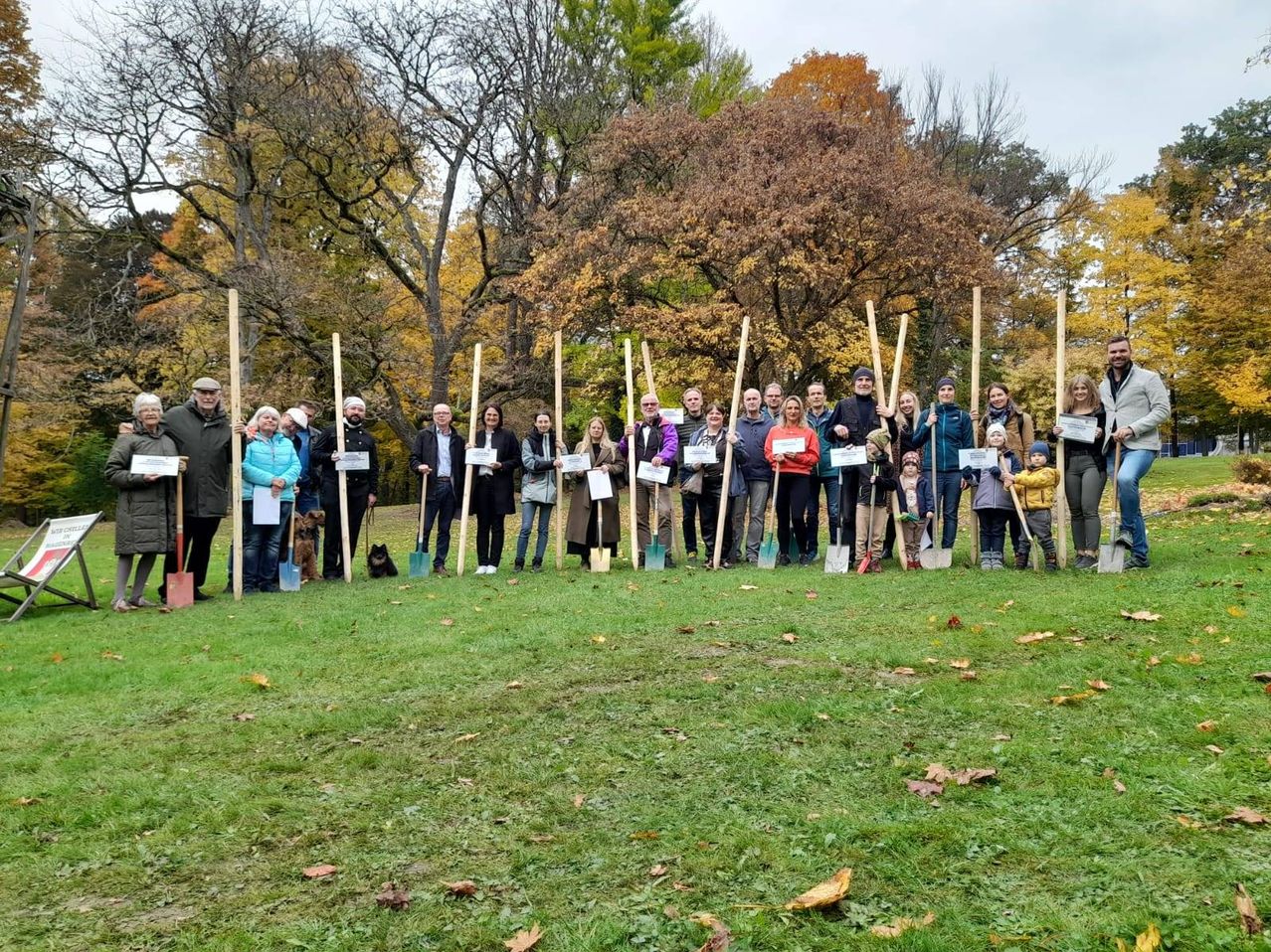 A group of people stands in a park with shovels, smiling and posing for a photo. Trees in fall colors form a backdrop.