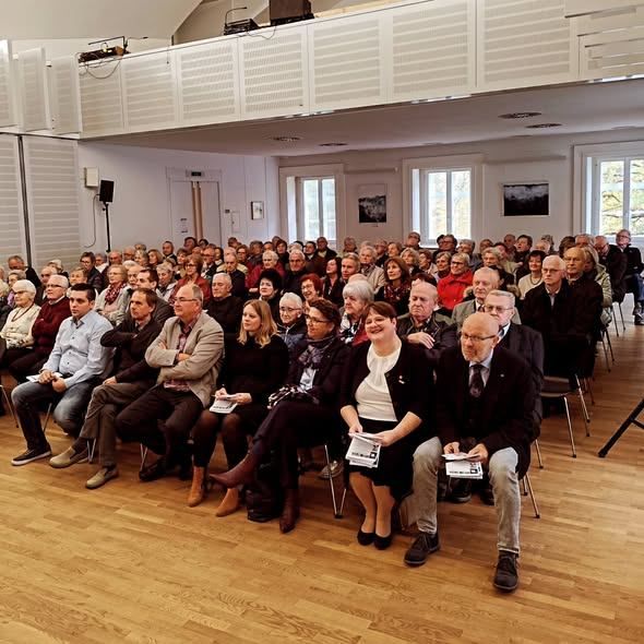 A large group of people are sitting in chairs in a room, likely attending a conference or meeting. They are wearing formal attire and some are holding papers. The room has wooden floors and white walls.