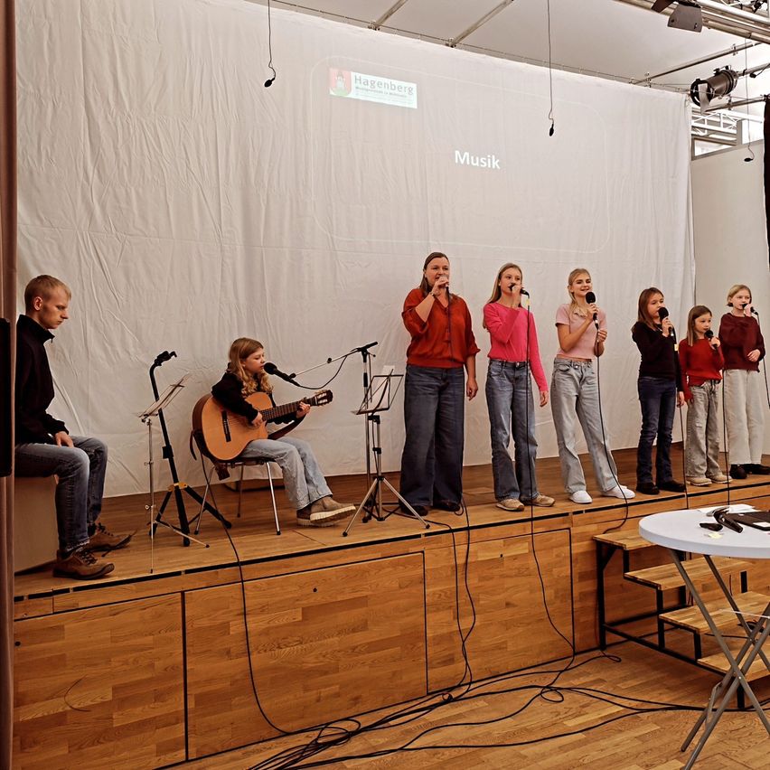 A group of children are on stage, performing with guitars and microphones. One boy plays guitar while seated. Other children stand, singing. Behind them, a white backdrop with 'Hagenberg' and 'Musik'.