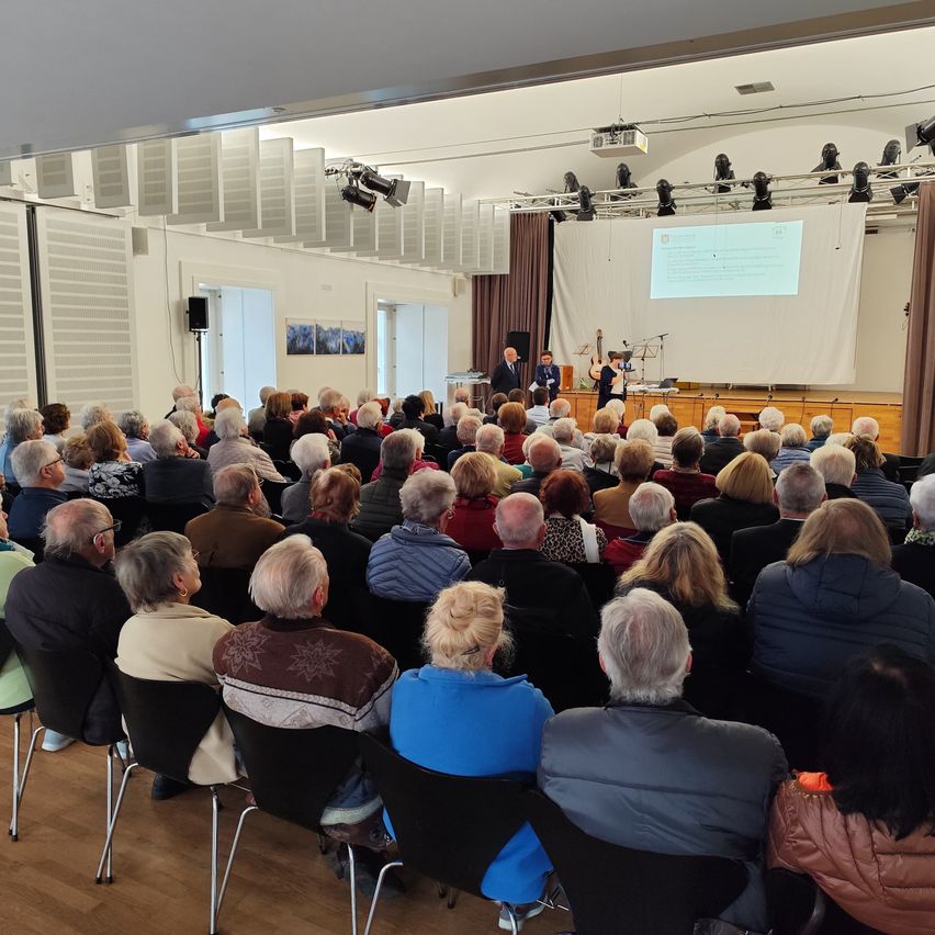 An auditorium filled with elderly people listening to a presentation with a projector screen and speakers on stage.