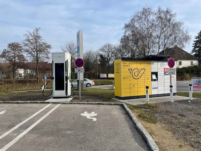 Ein Parkplatz mit einem gelben und weißen Postkasten und einer Elektroauto-Ladestation. Ein Auto ist am Straßenrand geparkt.