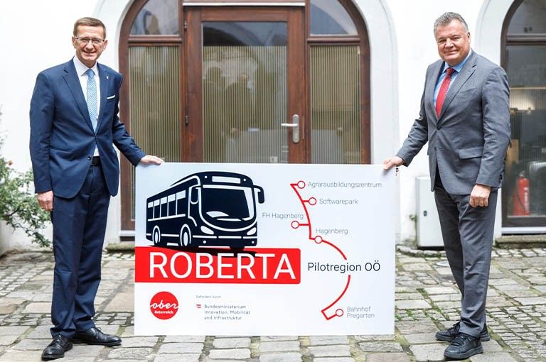 Zwei Männer stehen vor einem Schild mit einer Buslinienkarte, die 'ROBERTA Pilotregion Oö' betitelt ist. Beide Männer tragen Anzüge und Schuhe.