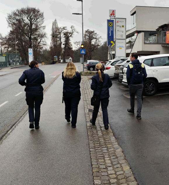 Four individuals in navy blue uniforms walk on the sidewalk near a parking lot with cars parked. There is a traffic light and road signs.