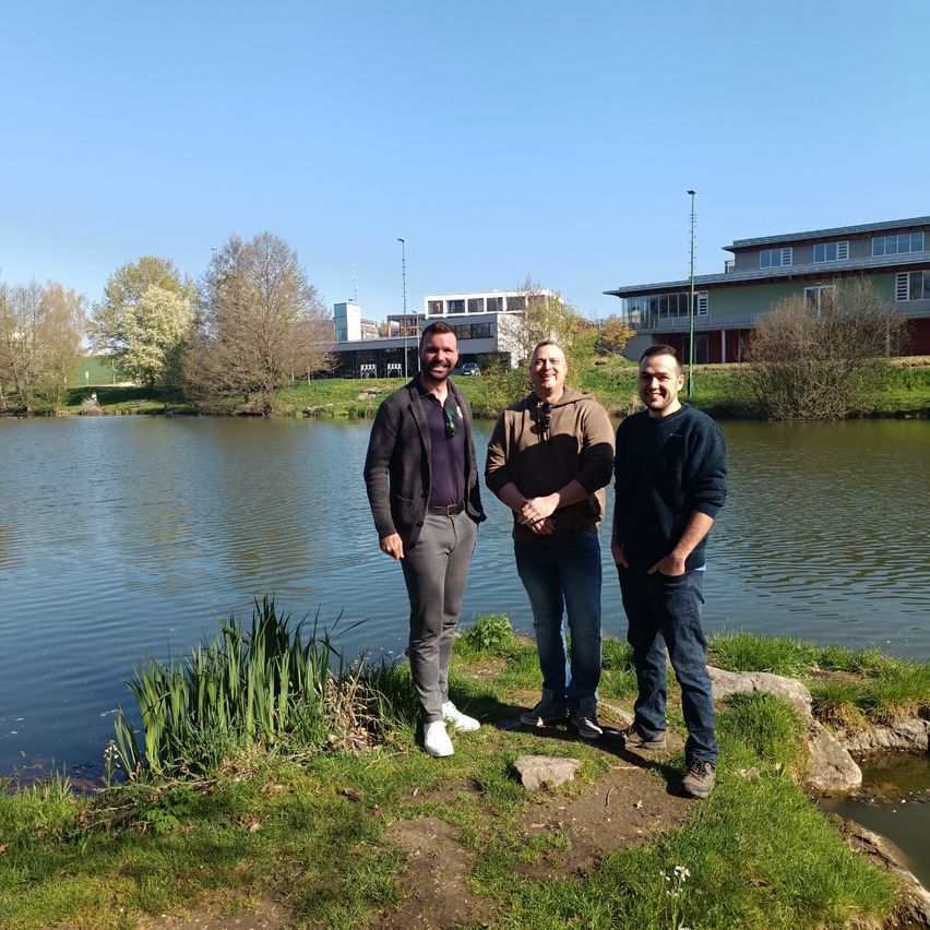 Drei Männer stehen am See, lächeln und posieren für ein Foto. Im Hintergrund sind Gebäude und Bäume zu sehen.