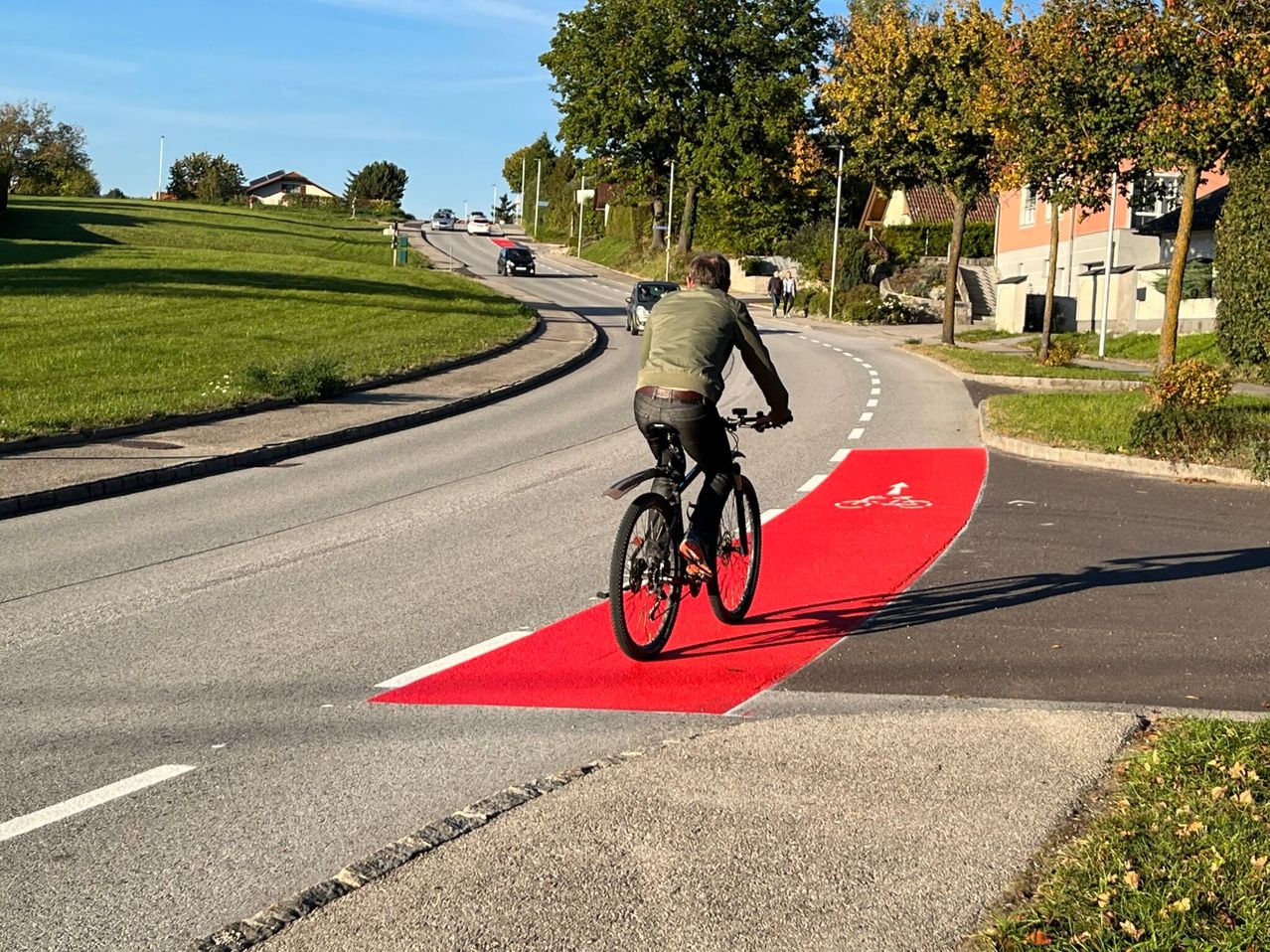 A man rides his bicycle on a red bike lane. Behind him, several cars drive on the road. On the right, houses and trees line the street.
