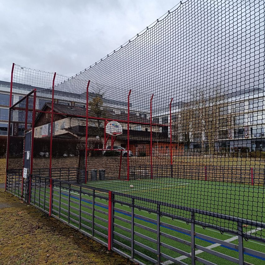 Ein eingezäunter Outdoor-Basketballplatz mit einem roten und schwarzen Netz. Der Platz ist leer, mit einem Ball auf dem Boden. Hinter dem Platz stehen Gebäude, und ein Auto ist geparkt.