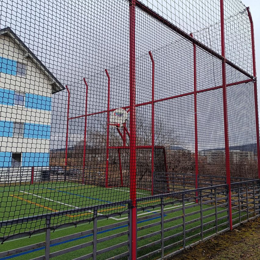 Ein Sportplatz ist von einem roten Zaun umgeben. Es gibt einen Basketballkorb mit Netz in der Mitte. Der Platz hat Kunstrasen.