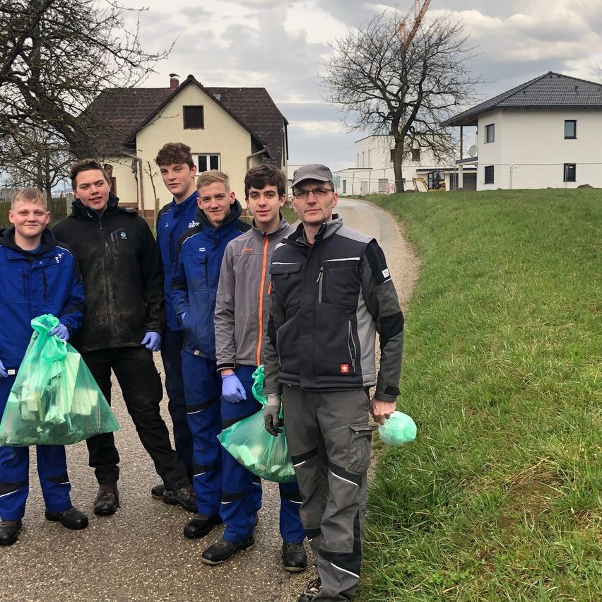 Sechs junge Männer in blauen und grauen Uniformen posieren mit Taschen in einem Wohngebiet, ein Haus und eine Baustelle im Hintergrund sichtbar.