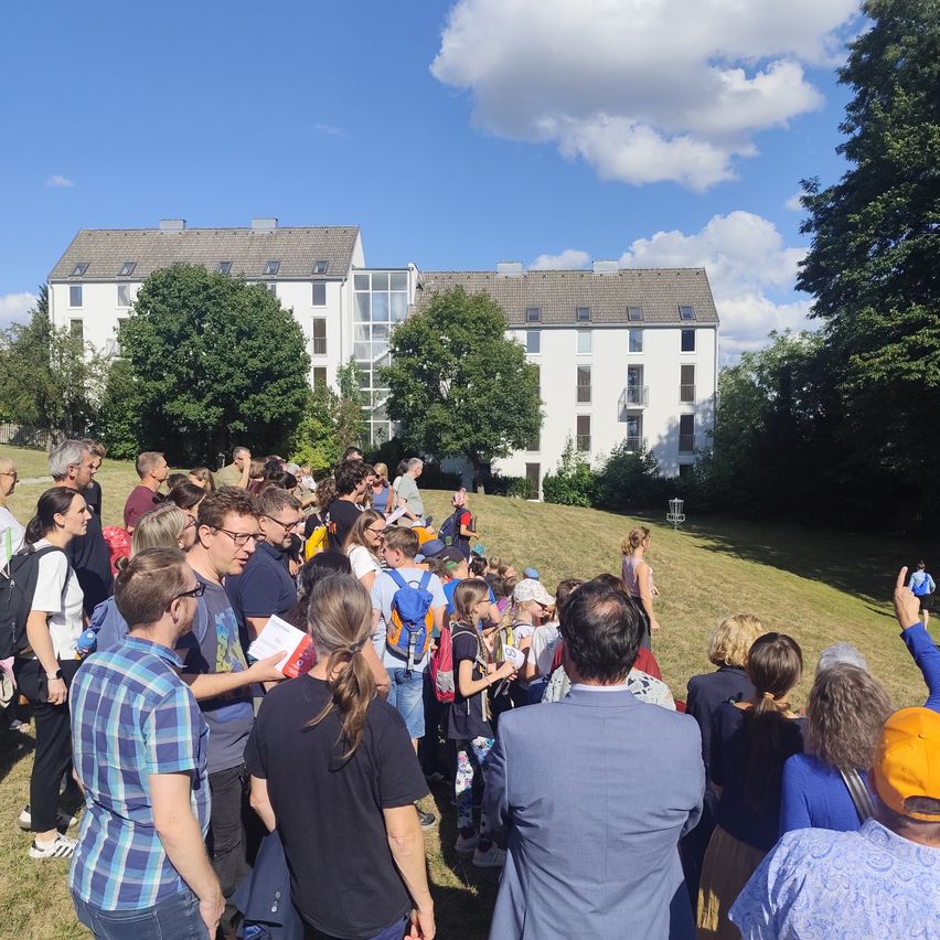 Eine Gruppe von Menschen versammelt sich auf einem Rasen vor einem großen weißen Gebäude. Einige tragen Brillen, Hüte und Turnschuhe. Eine Frau in einem weißen Shirt hält ein Stück Papier. Bäume und ein bewölkter Himmel sind im Hintergrund.