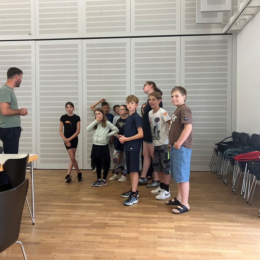 Eine Gruppe von Kindern und einem Mann stehen in einem Raum mit Holzboden und weißen Wänden. Der Mann spricht zu den Kindern.