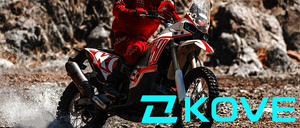 Ein Motorradfahrer in einem roten Anzug fährt ein Motorrad durch schlammiges Gelände. Das Motorrad hat eine große Windschutzscheibe und ist mit Schlamm bedeckt. Der Fahrer trägt einen Helm und Handschuhe.