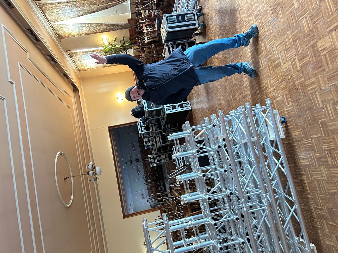 Ein Mann in einer blauen Jacke und Jeans hängt kopfüber von der Decke. Eine Lampe ist an der Wand und viele Metallkonstruktionen sind auf dem Boden.