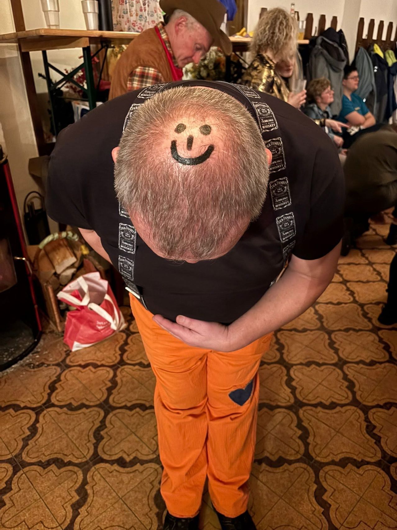 Eine Person mit einem lächelnden Gesicht auf dem kahlen Kopf, die sich in einem Raum mit gemustertem Boden bückt.