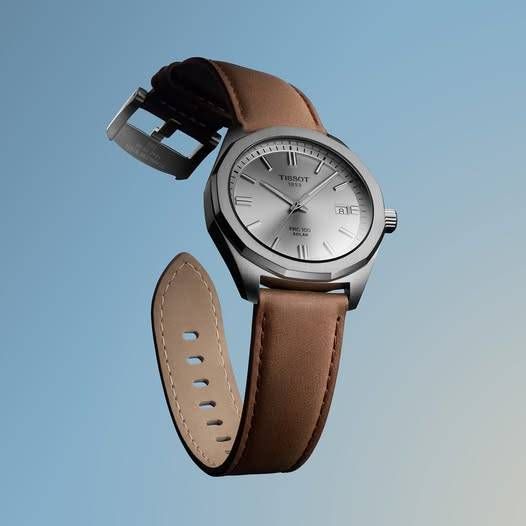 Eine silberne Tissot-Armbanduhr mit einem braunen Lederarmband schwebt am blauen Himmel. Das Zifferblatt zeigt römische Zahlen und den Markennamen.