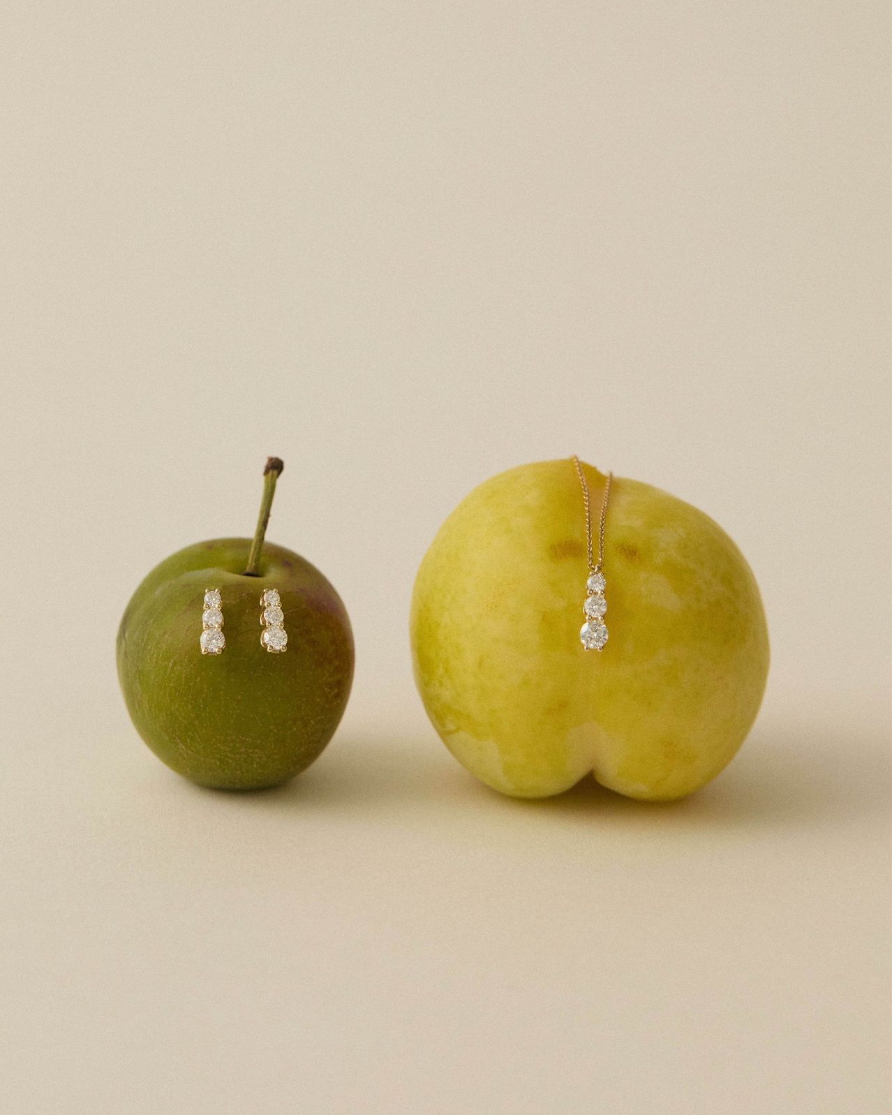 Zwei Äpfel, einer grün und einer gelb, werden mit Schmuck präsentiert. Der grüne Apfel hat zwei Diamantohrringe. Der gelbe Apfel hat eine Diamantkette.