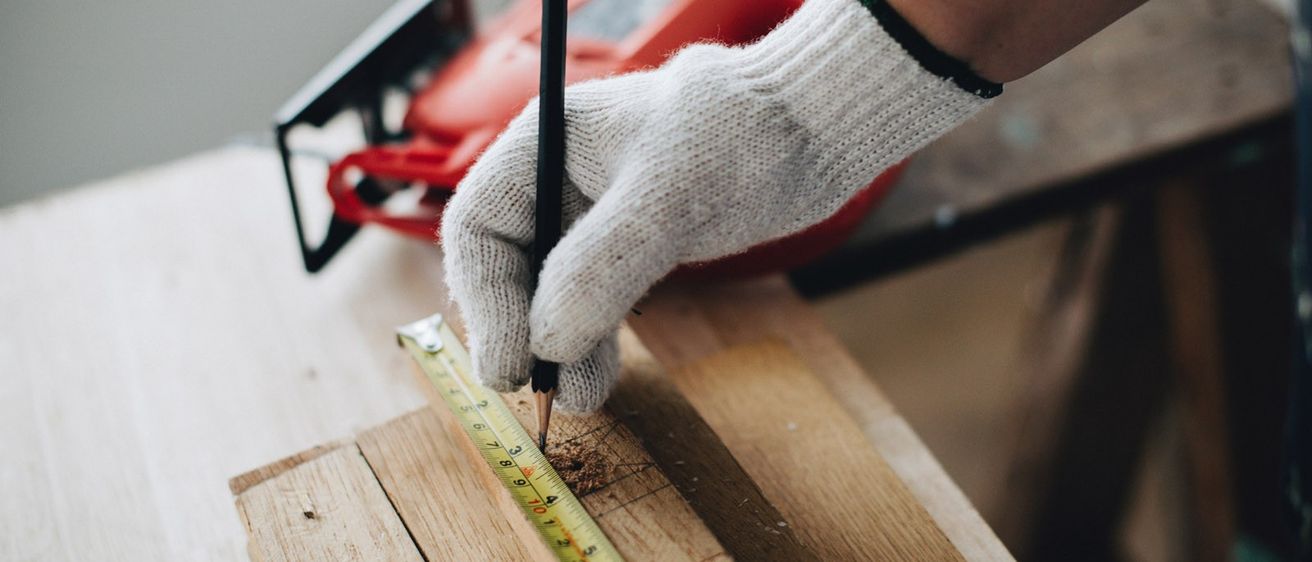 Eine Person in Handschuhen markiert eine Holzplatte mit einem Bleistift und misst sie mit einem Maßband.