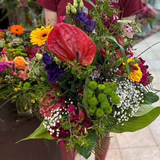 Ein farbenfroher Blumenstrauß mit roten, gelben, lila und grünen Blumen, gehalten von einer Person in einem roten T-Shirt.