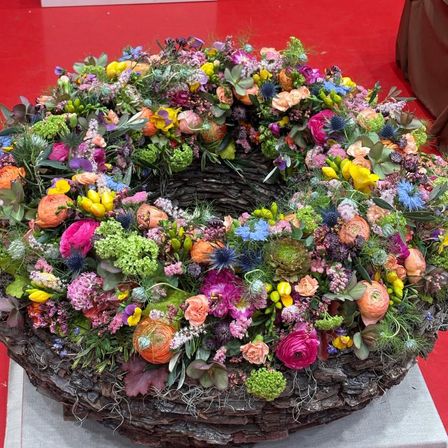 Eine große Blumenarrangement in einem Korb, bestehend aus einer Mischung von bunten Blumen und Grünpflanzen vor einem roten Hintergrund.