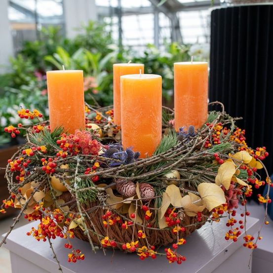 Eine dekorative Anordnung mit drei hohen orangefarbenen Kerzen, umgeben von einem Kranz aus Herbstblättern, Beeren und Tannenzapfen auf einer weißen Oberfläche.