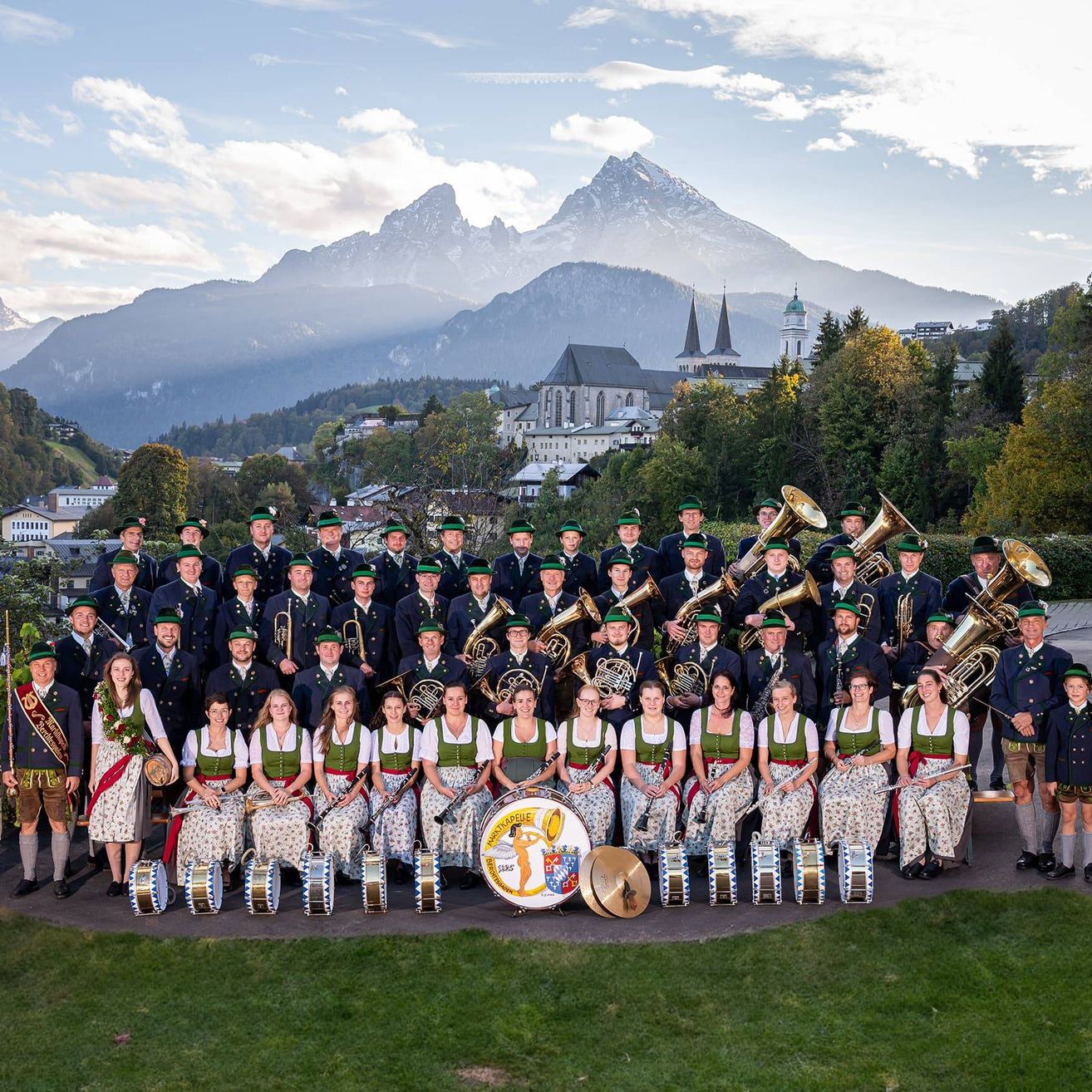 Eine große Gruppe von Musikern in traditioneller Kleidung posiert für ein Foto in einer malerischen Außenumgebung mit Bergen im Hintergrund.