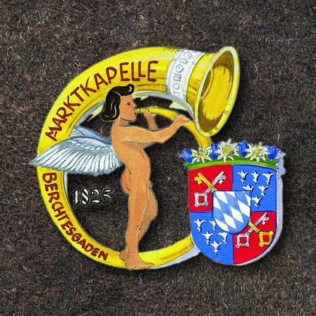 Ein Emblem mit einem Cherub, der in eine Trompete bläst, umgeben von Blumen. Das Emblem trägt den Text 'Marktkappelle' und das Jahr '1825'. Es zeigt auch ein Wappen mit Schlüsseln und Sternen.