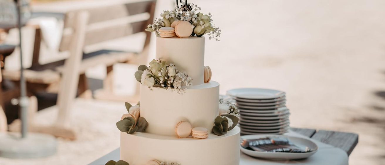 Ein dreistufiger Hochzeitskuchen mit weißer Glasur, verziert mit Macarons und Blumen, steht auf einem Holzsockel. Dahinter sind Teller und eine Bank zu sehen.