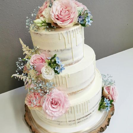 Ein dreistöckiger weißer Kuchen mit rosa Rosen und blauen Blumen, mit Zuckerguss überzogen, steht auf einem Holzsockel.
