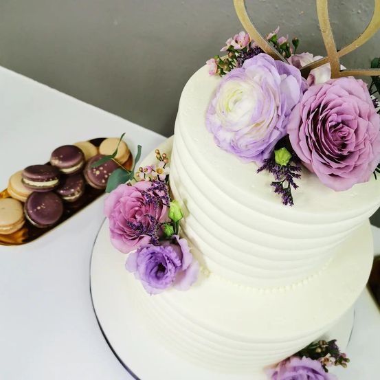 Ein dreistöckiger Hochzeitskuchen mit weißer Glasur, lila und weißen Blumen und einem herzförmigen goldenen Schild. Links steht ein Teller mit Schokoladen-Macarons.