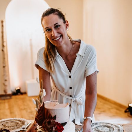 Eine Frau mit lächelndem Gesicht steht neben einem Kuchen. Sie trägt ein weißes Kleid und eine Halskette. Der Kuchen steht auf einem Holztisch.