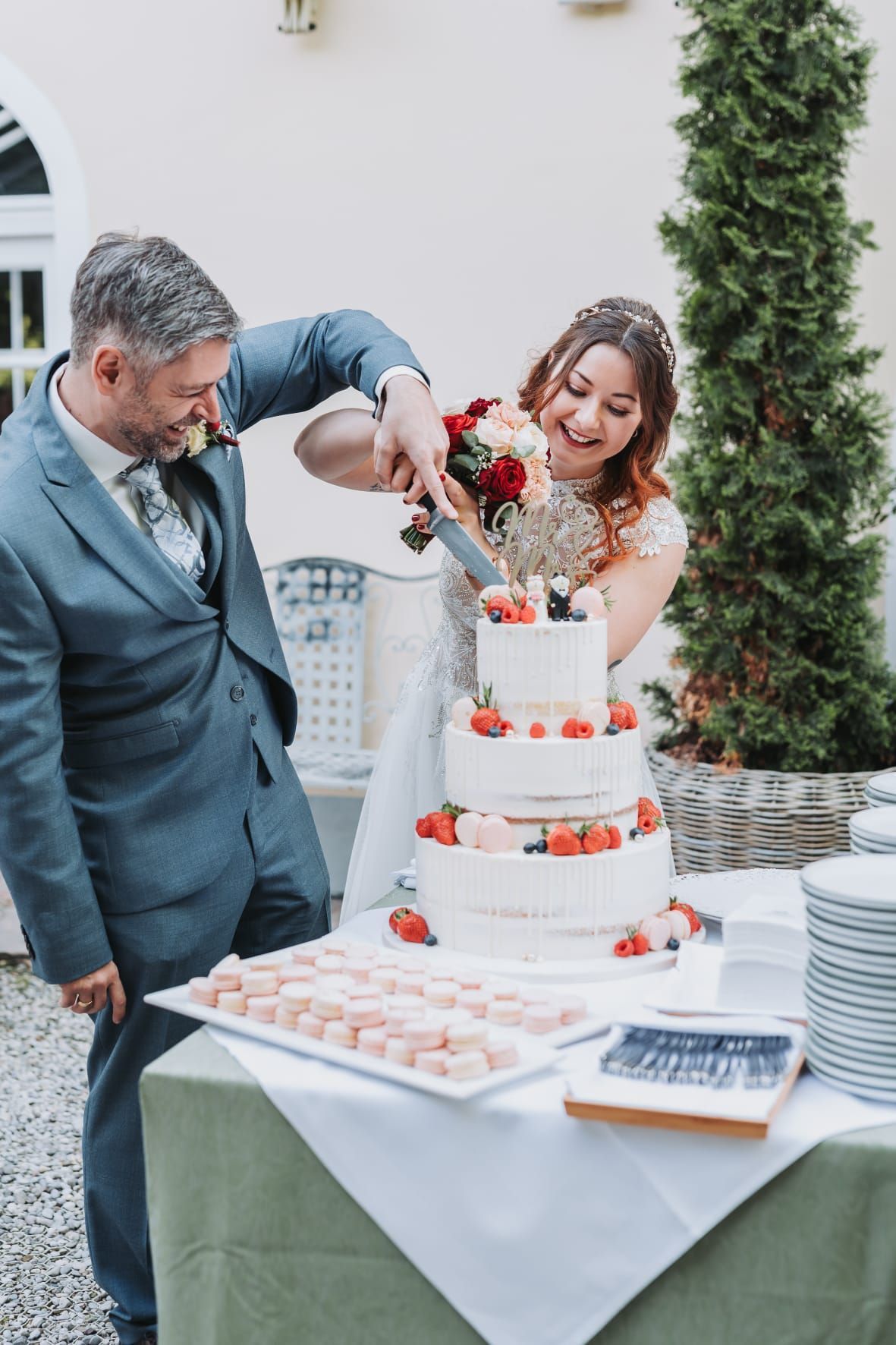 Ein frisch verheiratetes Paar schneidet in einen Hochzeitskuchen ein, der mit frischen Erdbeeren und Blaubeeren verziert ist, während sie lächeln und lachen.