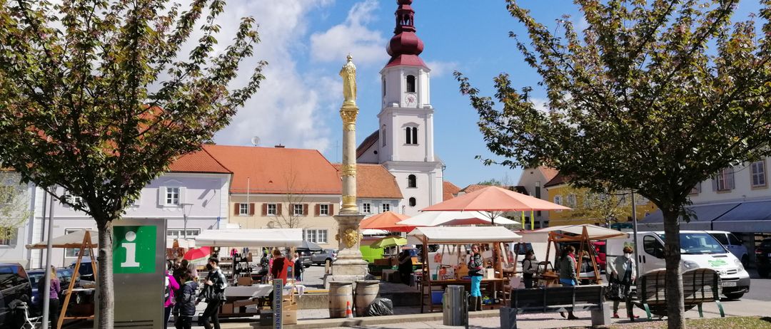 Eine Kirche mit rotem Dach steht inmitten eines Marktes mit mehreren Ständen unter Regenschirmen. Die Stände haben Menschen, die stöbern und Dinge kaufen. Bäume, Gras und eine Bank sind im Vordergrund. Ein Turm mit einer roten Kuppel und einer Uhr befindet sich auf der Kirche.