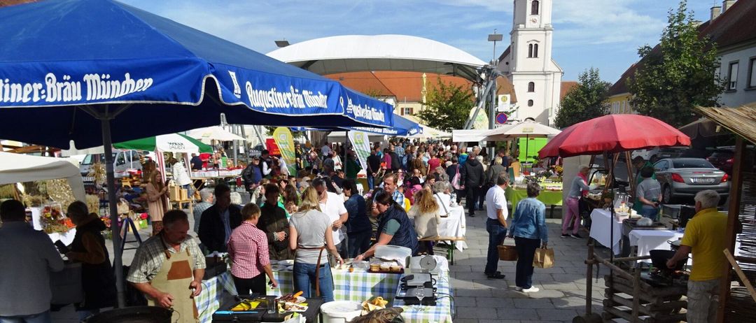 Ein Freiluftmarkt mit vielen Leuten. Stände sind mit blauen und weißen Zelten abgedeckt. Ein Turm ist im Hintergrund.