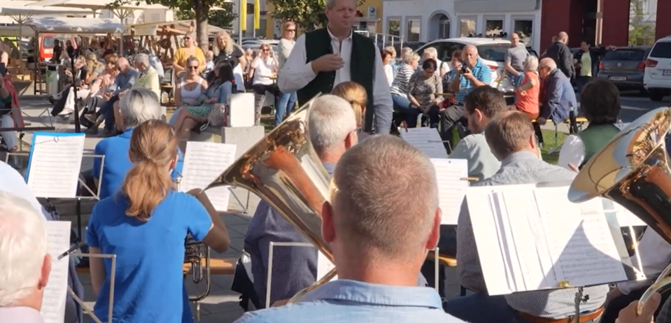 Ein Orchester spielt auf einem öffentlichen Platz mit einem großen Publikum, unter einem Baum und in der Nähe von Gebäuden.