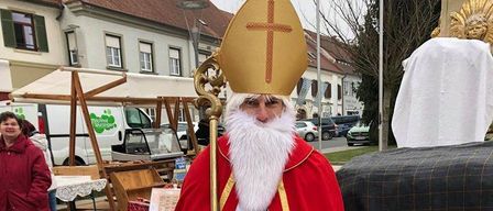 Ein Mann als Bischof gekleidet steht vor einer Bank und hält ein Buch in der Hand, während eine Frau in der Nähe eines Marktstandes steht und ein Lieferwagen im Hintergrund zu sehen ist.