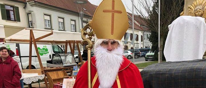 Ein Mann als Bischof gekleidet steht vor einer Bank und hält ein Buch in der Hand, während eine Frau in der Nähe eines Marktstandes steht und ein Lieferwagen im Hintergrund zu sehen ist.