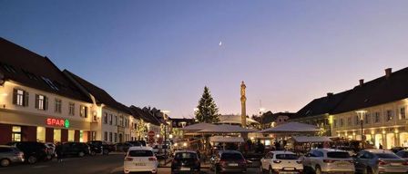 Ein Stadtplatz in der Dämmerung mit einem Halbmond, einem Weihnachtsbaum und mehreren geparkten Autos vor Gebäuden. Ein Gebäude hat ein Spar-Schild.