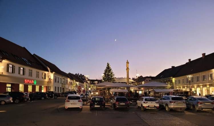 Ein Stadtplatz in der Dämmerung mit einem Halbmond, einem Weihnachtsbaum und mehreren geparkten Autos vor Gebäuden. Ein Gebäude hat ein Spar-Schild.